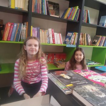 Biblioteka szkolna. Dwie uśmiechnięte dziewczynki siedzą przy biurku. Na biurku leżą książki. Z tyłu widać półki z książkami.