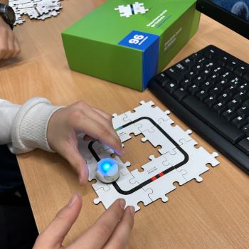 Uczeń trzyma w rękach świecącego na niebiesko ozobota. Na stoliku leży klawiatura, pudełko i kody ułożone w trasę w kształcie kwadratu.