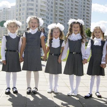 Dziewczynki w odświętnych ubraniach stoją przed wysokim budynkiem. Są uśmiechnięte, trzymają się za ręce i wyglądają jakby nie mogły doczekać się pójścia do szkoły.